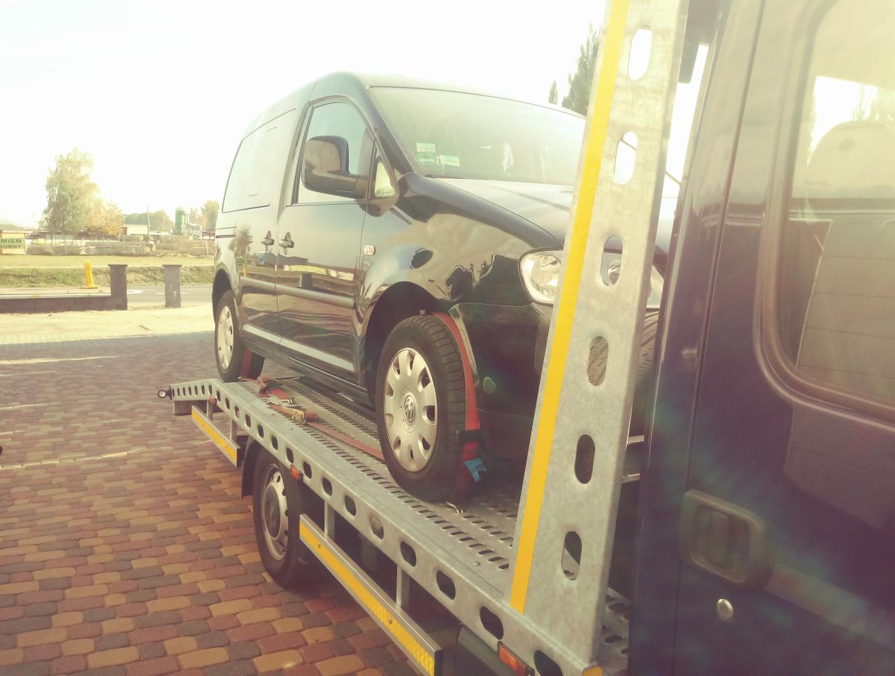Czarny Volkswagen Caddy zabezpieczony pasami transportowymi na platformie lawety, widok z boku, w tle kostka brukowa i zieleń.