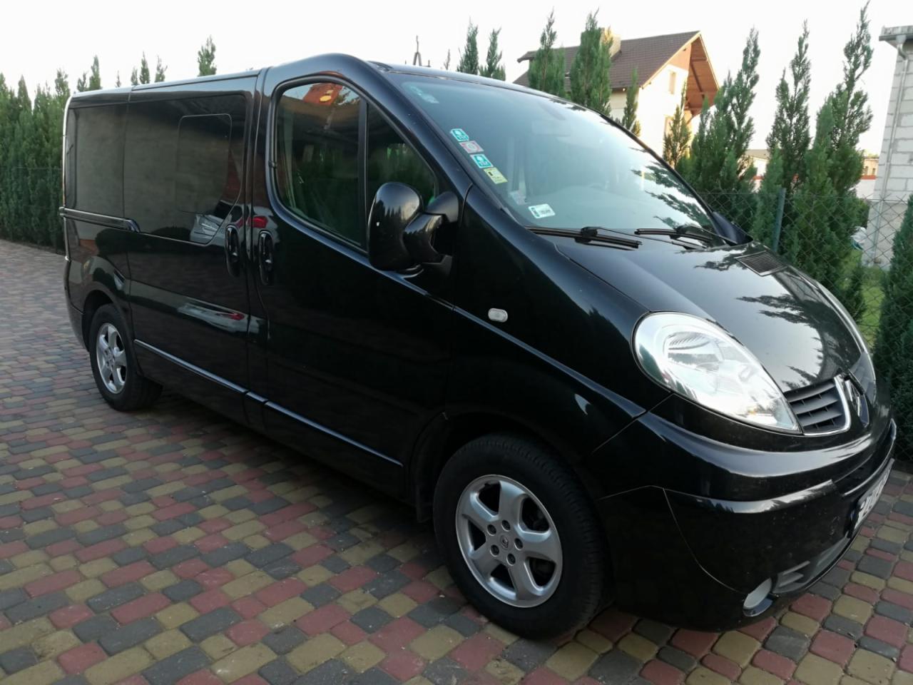 Czarny bus Renault Trafic z przyciemnianymi szybami, zaparkowany na kostce brukowej przed ogrodzeniem z tujami i domem w tle.