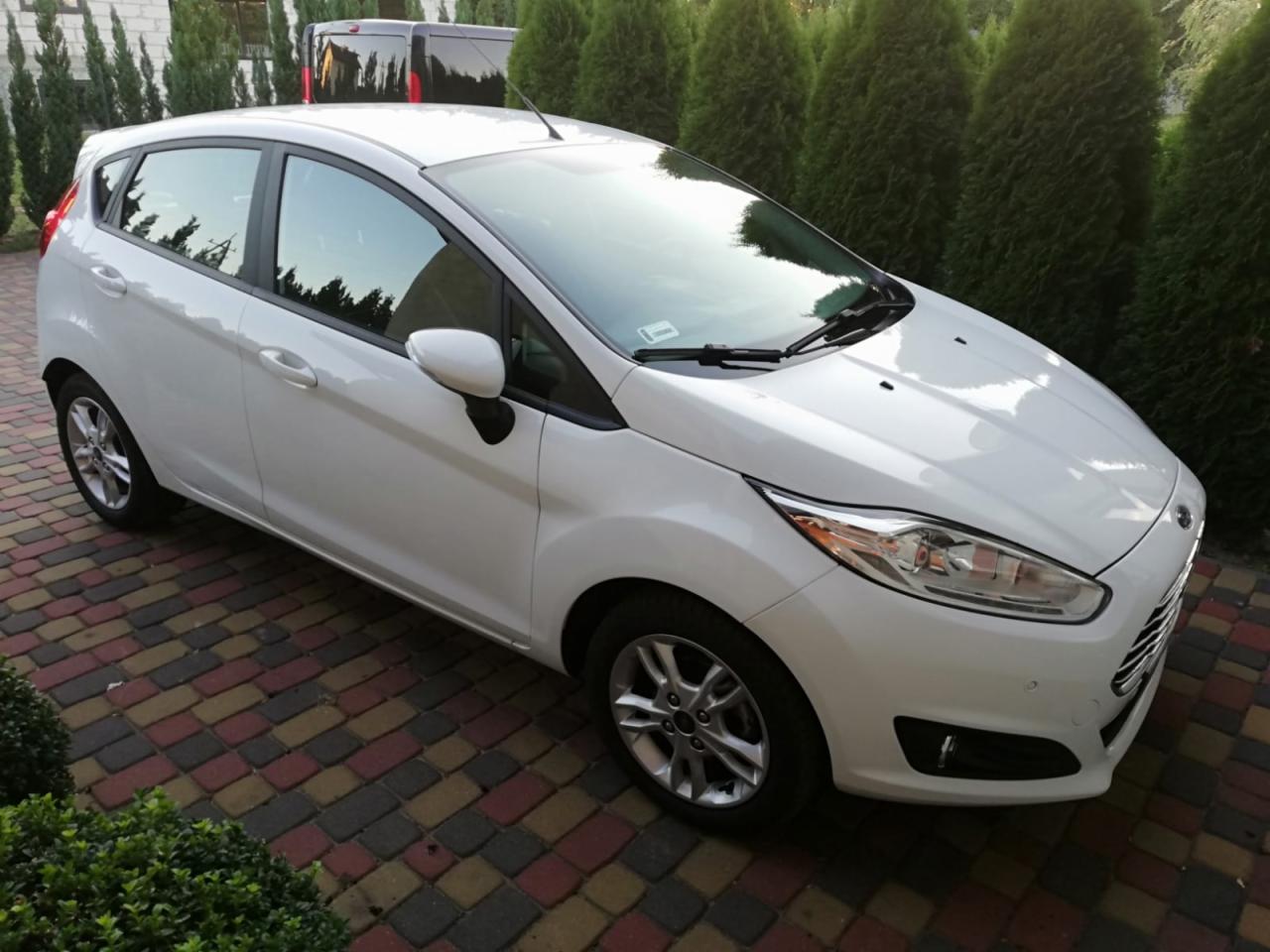 Biały Ford Fiesta hatchback zaparkowany na kolorowej kostce brukowej, tło stanowią zielone tuje i ciemny samochód dostawczy.