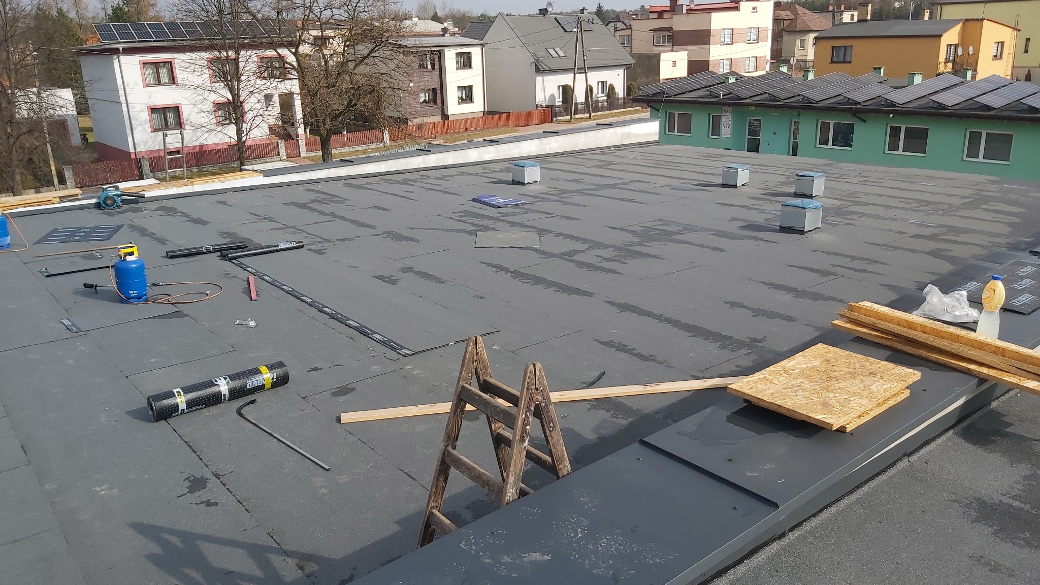 Rozległy, płaski dach pokryty czarną papą termozgrzewalną w trakcie prac. Widoczne narzędzia: butla gazowa, rolka papy, drabina drewniana, deski, płyty OSB. W tle budynki mieszkalne i panele...