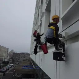 Dwóch alpinistów na linach maluje elewację budynku w miejskiej scenerii; jeden z nich, w żółtym kasku, maluje białą farbą, drugi w czerwonym kasku pokazuje coś ręką.