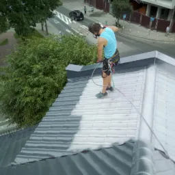 Pracownik w uprzęży, z maską na twarzy, używa pistoletu natryskowego do pokrywania dachu z blachodachówki szarą farbą. Część dachu jest już pomalowana, widoczny kontrast między starą a nową powłoką...