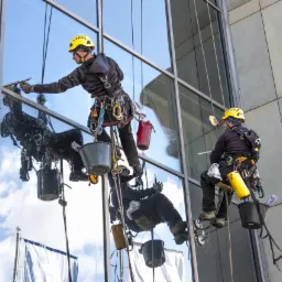 Dwóch mężczyzn w uprzężach i kaskach czyści szklaną elewację wysokiego budynku, używając ściągaczek i wiader zawieszonych na linach.