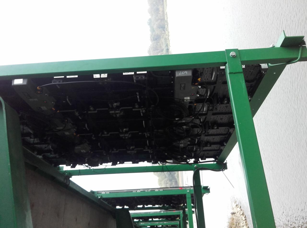 Widok od dołu na konstrukcję metalową w kolorze zielonym, prawdopodobnie rusztowanie, z zamontowanymi modułami elektronicznymi i okablowaniem; w tle widać wodę i drzewa.