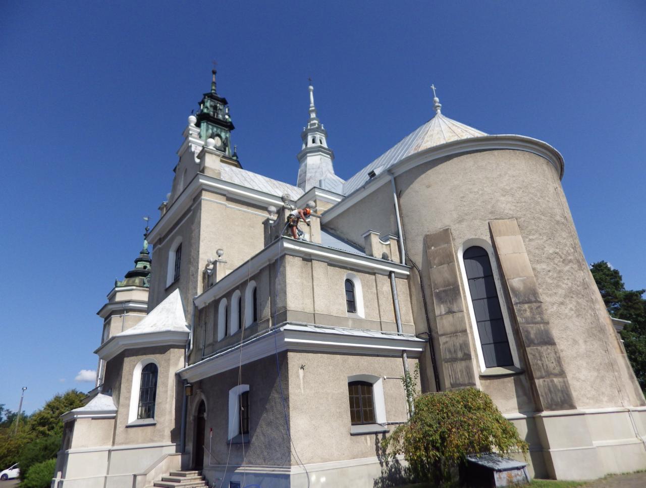 Dwóch pracowników na wysokościach, zabezpieczonych linami, wykonuje prace na elewacji zabytkowego kościoła z wieżami i kopułą, widok z dołu na tle błękitnego nieba.