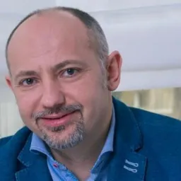 Portret uśmiechniętego, łysiejącego mężczyzny z brodą w niebieskiej marynarce na tle okna z białymi roletami i ramami, skierowany lekko w prawo.