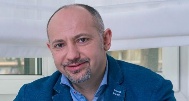 Portret uśmiechniętego, łysiejącego mężczyzny z brodą w niebieskiej marynarce na tle okna z białymi roletami i ramami, skierowany lekko w prawo.