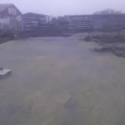 Przygotowywanie podłoża pod kostkę brukową: pracownik wyrównuje piasek na placu budowy, widoczne narzędzia i materiały na palecie, w tle budynki gospodarcze.