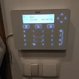 Biały panel sterowania alarmem firmy Risco z wyświetlaczem LCD pokazującym godzinę i datę, zamontowany na ścianie nad potrójnym włącznikiem światła.