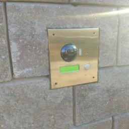 Złoty panel wideodomofonu z zielonym wyświetlaczem zamontowany na elewacji z kamienia łupanego, widoczne odbicie otoczenia w obiektywie kamery.