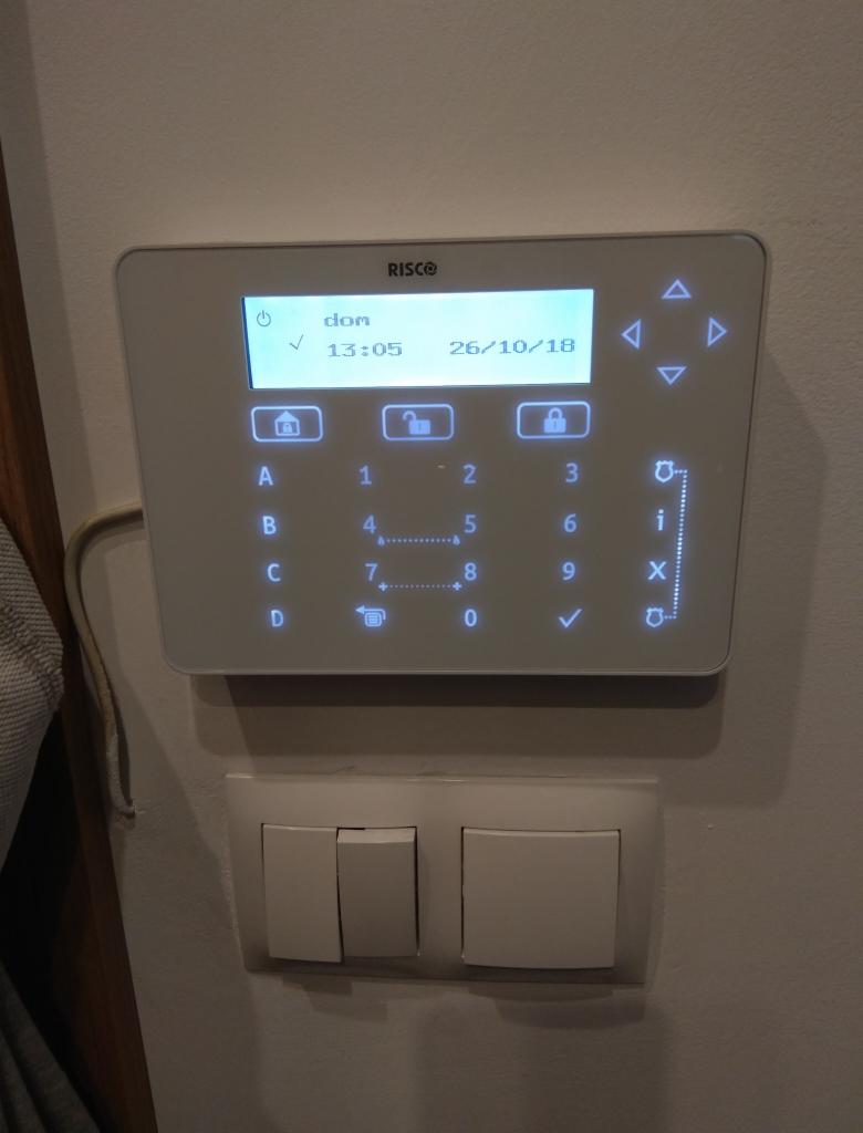 Biały panel sterowania alarmem firmy Risco z wyświetlaczem LCD pokazującym godzinę i datę, zamontowany na ścianie nad potrójnym włącznikiem światła.