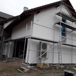 Marcin Turkiewicz DRAGON - Dom jednorodzinny w trakcie prac elewacyjnych: świeżo nałożony biały tynk, widoczne rusztowania, klimatyzator na elewacji, pracownik wewnątrz budynku.