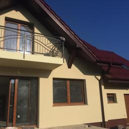 Marcin Turkiewicz DRAGON - Żółta elewacja domu jednorodzinnego z brązowym dachem i balkonem z metalową balustradą, widok z zewnątrz w słoneczny dzień.