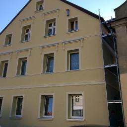 Marcin Turkiewicz DRAGON - Odnowiona elewacja kamienicy w odcieniach beżu z dekoracyjnymi elementami wokół okien, częściowo zasłonięta przez metalowe rusztowanie.