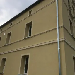 Odnowiona elewacja budynku z beżowym tynkiem strukturalnym, ozdobnymi gzymsami i nową rynną spustową.