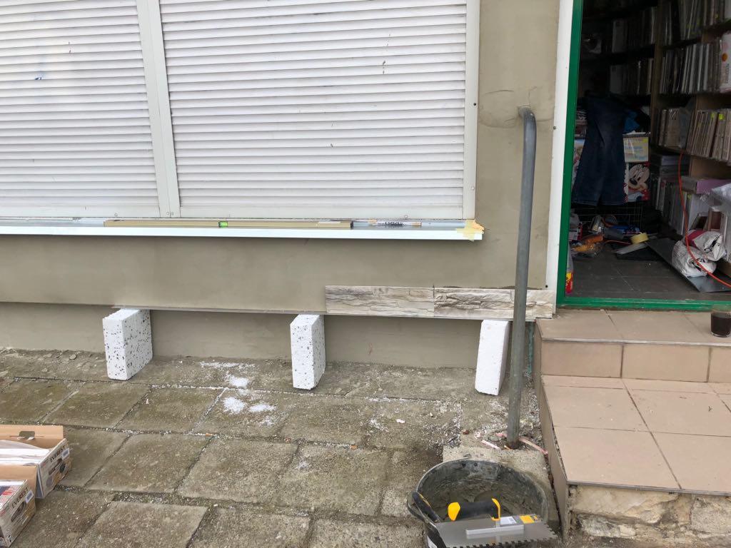 Fragment elewacji budynku z częściowo nałożonymi płytkami imitującymi kamień, podparte bloczkami betonowymi, widoczny poziomica i wiadro z narzędziami.