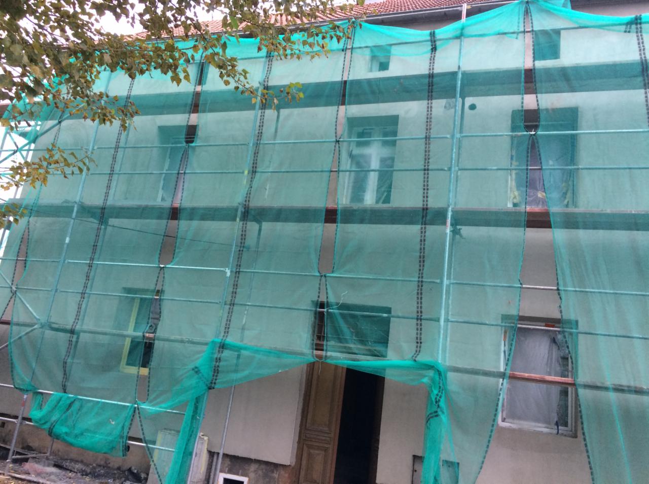 Dom jednorodzinny w trakcie prac elewacyjnych, osłonięty siatką ochronną na rusztowaniu, widoczne okna zabezpieczone folią.