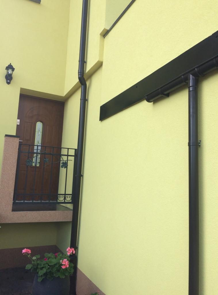 Ściana budynku w jasnym odcieniu z zamontowanym systemem rynnowym w kolorze czarnym, widoczny fragment wejścia z drewnianymi drzwiami i dekoracyjną balustradą oraz donica z kwitnącymi pelargoniami.