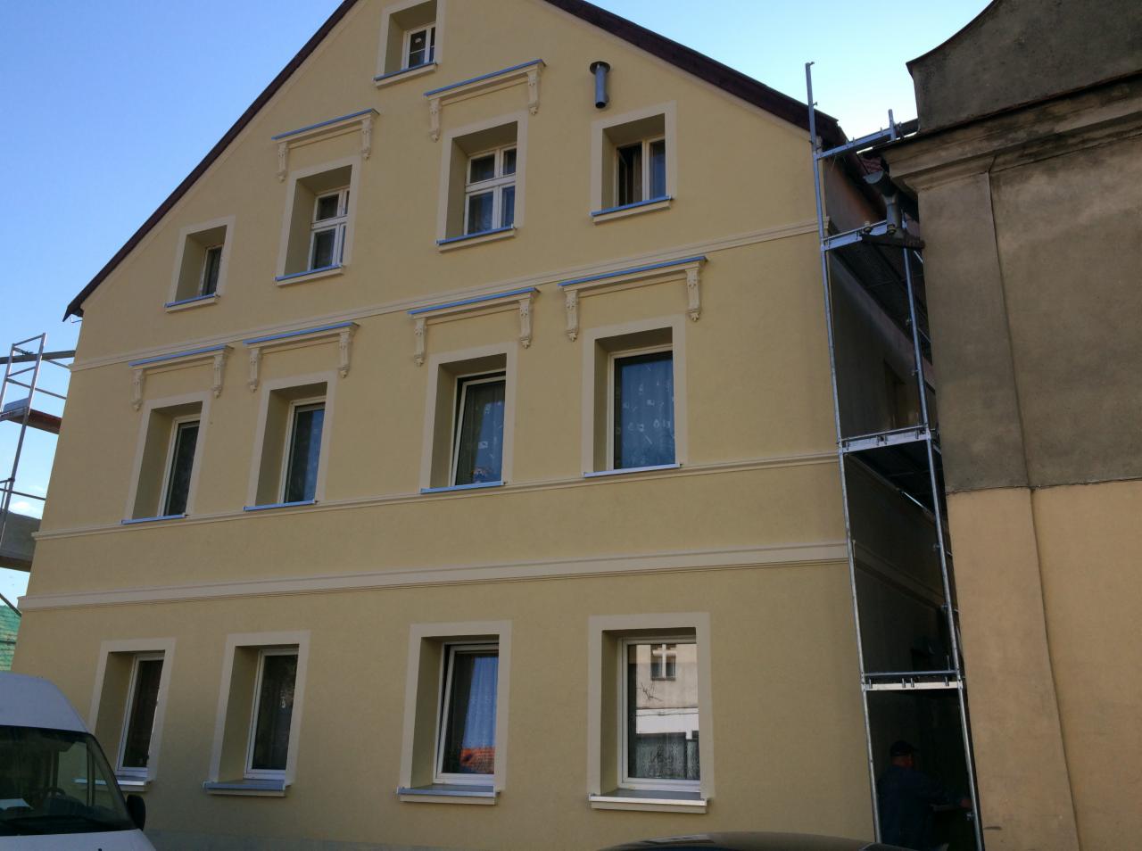 Odnowiona elewacja kamienicy w odcieniach beżu z dekoracyjnymi elementami wokół okien, częściowo zasłonięta przez metalowe rusztowanie.
