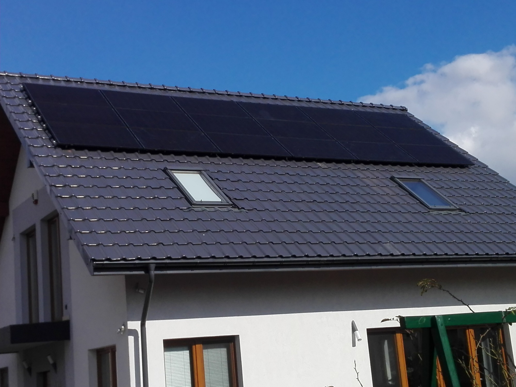 Czarny panel fotowoltaiczny zamontowany na grafitowym dachu z dachówki ceramicznej nowoczesnego domu jednorodzinnego z oknami dachowymi, na tle błękitnego nieba z chmurami.