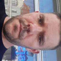 Portret mężczyzny z krótką brodą i ciemnymi włosami, w czarnej koszulce, z budynkiem w tle, obrócony o 90 stopni.