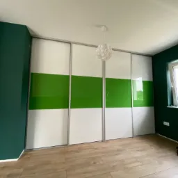 Czterodrzwiowa szafa przesuwna z białymi i zielonymi panelami, widoczna w pokoju z drewnopodobną podłogą i ścianami w kolorze butelkowej zieleni.