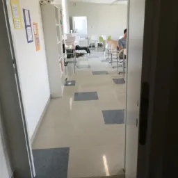 Widok zza uchylonych drzwi na jasne pomieszczenie biurowe z biurkami, krzesłami i regałami, z widoczną osobą siedzącą przy jednym z biurek.