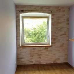 Pokój po remoncie: ściana z imitacją kamienia, okno z widokiem na zieleń, drewnopodobna podłoga, pomalowana na fioletowo ściana z półką.