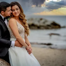 Para młoda w objęciach na plaży o zachodzie słońca, panna młoda w sukni z koronkową górą, pan młody w czarnym smokingu z muchą, w tle morze i chmury z promieniami słońca.