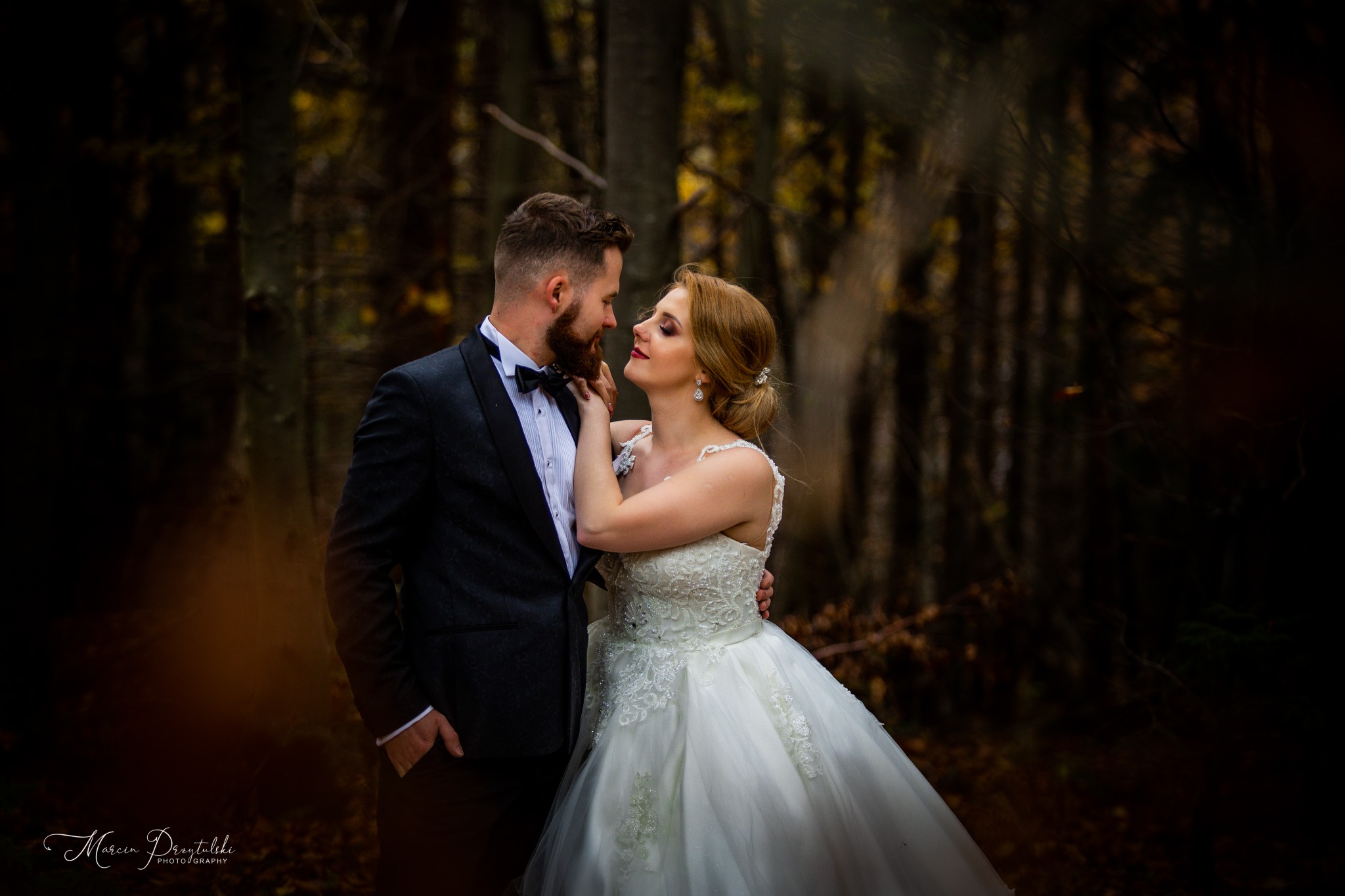 Para młoda w lesie; panna młoda dotyka twarzy pana młodego, oboje patrzą sobie w oczy, tło stanowią drzewa z jesiennymi liśćmi.