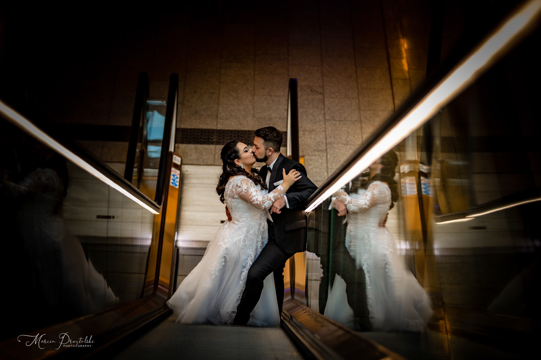 Pocałunek nowożeńców na schodach ruchomych, panna młoda w koronkowej sukni z długim rękawem, pan młody w czarnym smokingu, odbicie w szkle balustrady.