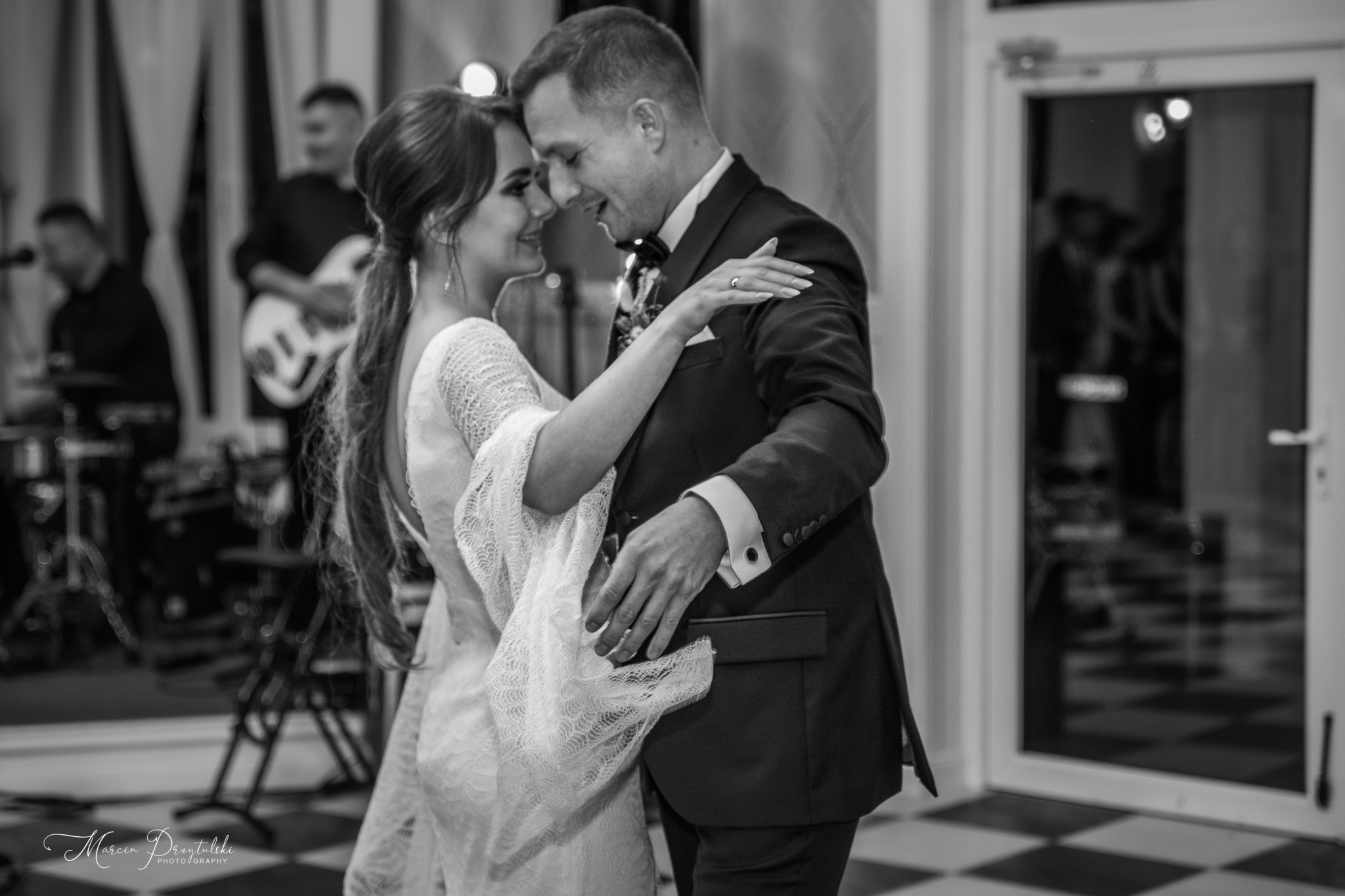 Czarno-białe zdjęcie pary młodej w tańcu, z zespołem muzycznym w tle, podczas wesela na sali z szachownicą na podłodze.