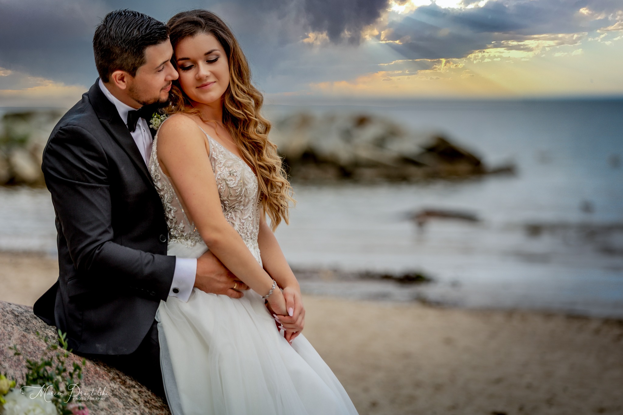 Para młoda w objęciach na plaży o zachodzie słońca, panna młoda w sukni z koronkową górą, pan młody w czarnym smokingu z muchą, w tle morze i chmury z promieniami słońca.