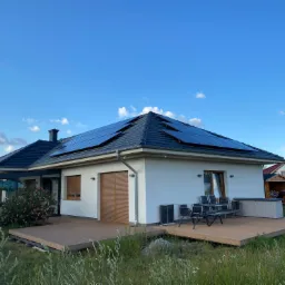 Biały dom jednorodzinny z ciemną dachówką i panelami słonecznymi na dachu, otoczony drewnianym tarasem i zielenią, na tle błękitnego nieba z chmurami.