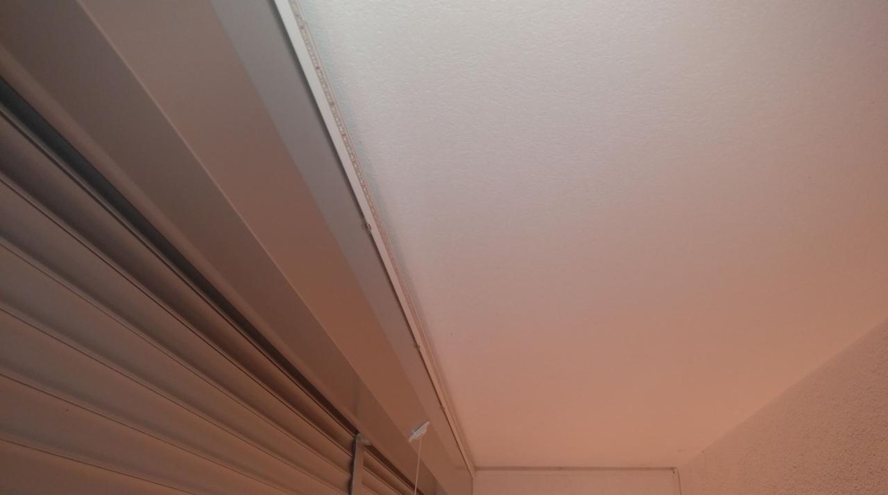 Widok na biały sufit z zainstalowanym oświetleniem LED wzdłuż krawędzi oraz fragmentem szarych żaluzji okiennych po lewej stronie.