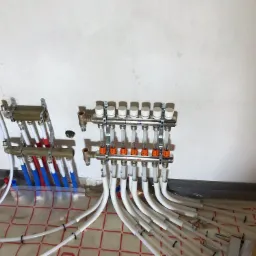 Rozdzielacz ogrzewania podłogowego z białymi rurkami, zamontowany na ścianie w surowym stanie, z widoczną folią izolacyjną na podłodze z czerwoną siatką. Część rurek w kolorze niebieskim...