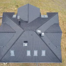 Widok z góry na nowy dach z ciemnej dachówki na domu w trakcie budowy, widoczny podnośnik koszowy i materiały budowlane.