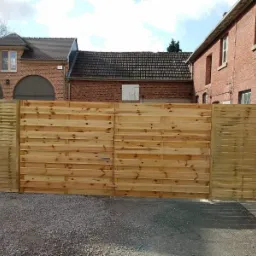 Nowo postawione drewniane ogrodzenie panelowe z surowego drewna na tle ceglanych budynków z dachówką ceramiczną, widoczne z zewnątrz posesji.