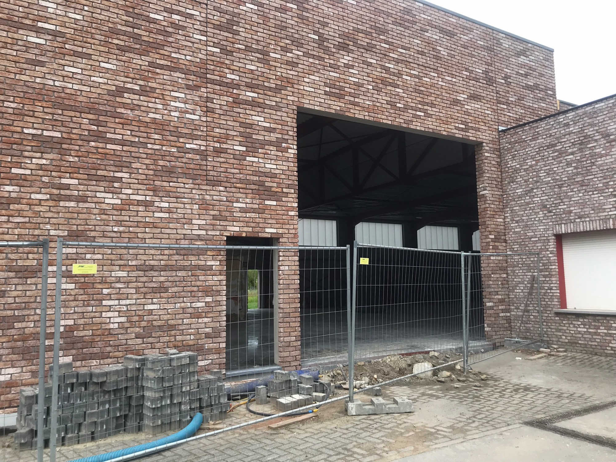 Wznoszony budynek z cegły klinkierowej, widoczne otwory okienne i drzwiowe, teren budowy zabezpieczony barierkami, przygotowane kostki brukowe.