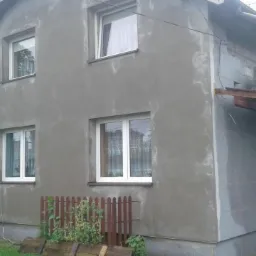 Dom jednorodzinny w trakcie tynkowania elewacji, widoczne ślady po nałożonym tynku, białe okna i fragment rusztowania z lewej strony.