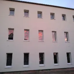 Trzypiętrowy budynek z jasną elewacją i regularnie rozmieszczonymi oknami, w których odbija się zachodzące słońce i drzewa.