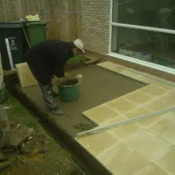 Pracownik w czarnej kurtce i jasnej czapce układa beżowe płyty chodnikowe na zaprawie, widoczna betoniarka i okno domu z cegły w tle.