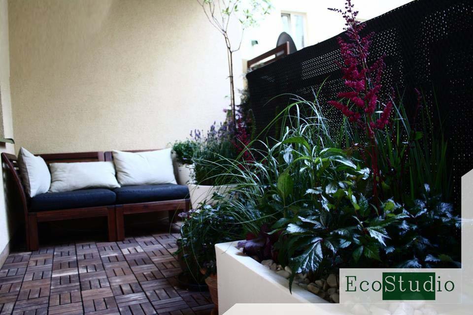 Kompaktowy ogród na balkonie z drewnianą podłogą, sofą z poduszkami i roślinami w białych donicach, osłonięty czarną siatką.