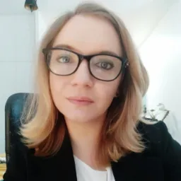 Portret młodej kobiety w okularach i czarnej marynarce, z delikatnym uśmiechem, siedzącej na krześle biurowym, z rośliną w tle.