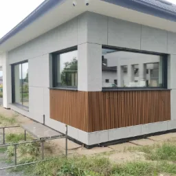 Nowoczesny dom z szarą elewacją panelową i drewnianymi akcentami, widoczne rusztowanie i teren w trakcie prac wykończeniowych.