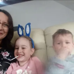 Kobieta w okularach i dwoje dzieci siedzą na jasnej kanapie: dziewczynka z lizakiem i niebieskimi uszami królika, chłopiec z ręką w buzi.