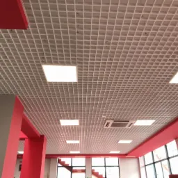 Kasetonowy sufit podwieszany z wbudowanymi panelami LED, widoczne elementy instalacji wentylacyjnej, czerwone filary i fragment schodów w tle.