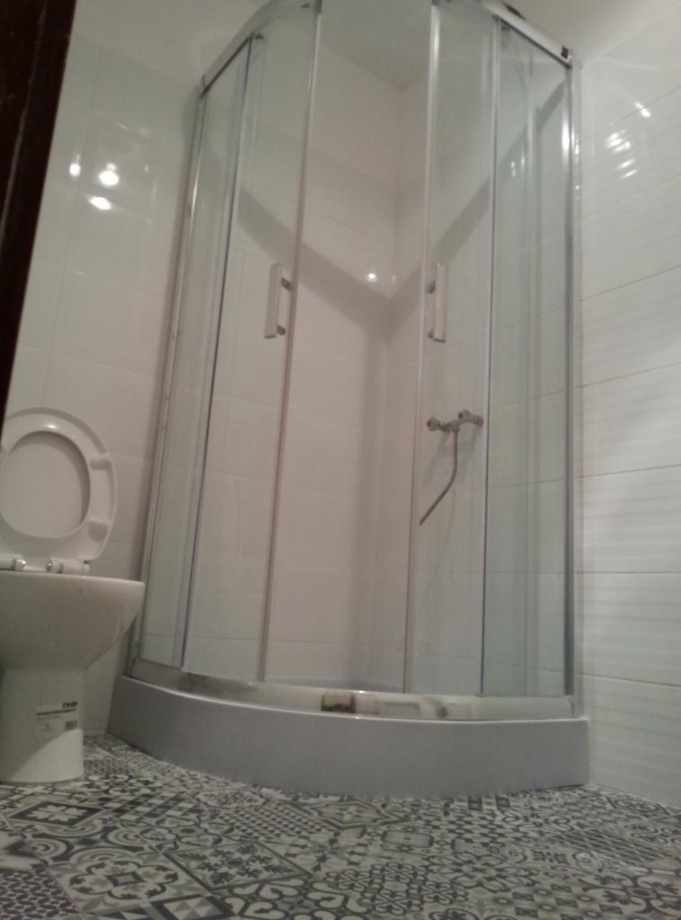 Narożna kabina prysznicowa z przezroczystymi drzwiami i chromowanymi uchwytami, widoczna obok toaleta z otwartą klapą, wzorzysta podłoga i białe płytki na ścianach.