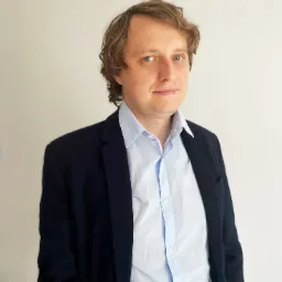 Portret mężczyzny w niebieskiej koszuli i granatowej marynarce na jasnym tle, z delikatnym uśmiechem i spojrzeniem w obiektyw.