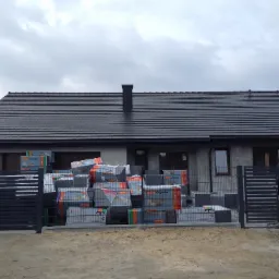 Nowoczesny dom jednorodzinny z ciemną dachówką, stertą opakowań termoizolacyjnych przed ogrodzeniem, widoczne okna i komin, pochmurne niebo.